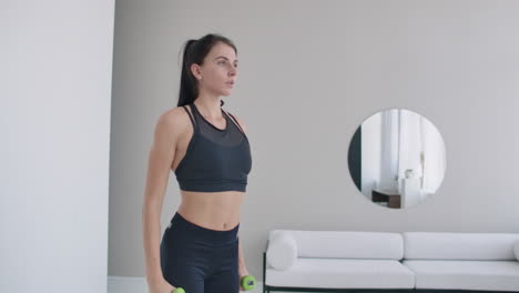 A-woman-raises-her-arms-with-dumbbells-doing-a-shoulder-exercise-in-her-light-white-apartment-against-a-sofa-and-window.